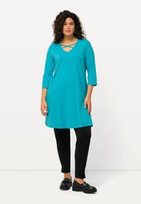 Türkise Tunika mit V-Ausschnitt und Kreuzdesign, kombiniert mit schwarzen Leggings. Der Stoff ist weich und leicht, mit ¾-Ärmeln und lockerer Passform.