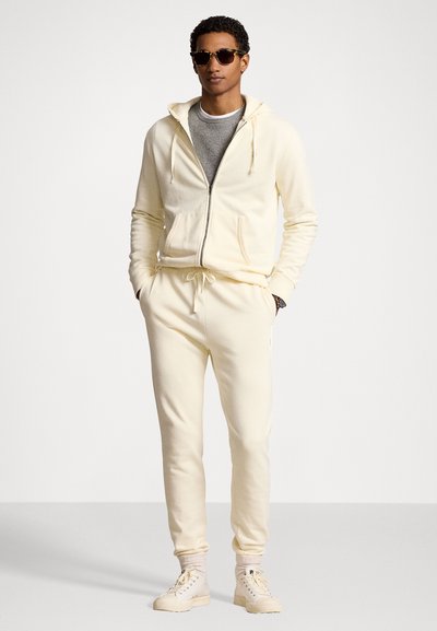 Lys gul hættejakke med lynlås, snore og frontlommer, parret med matchende sweatpants. Båret med en grå T-shirt og solbriller.