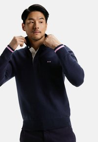 Pull-over marin avec col à boutons blancs, poignets rayés rose et marine, et un subtil détail de logo sur la poitrine. Texture en maille douce.