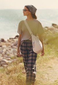 Femme portant des lunettes de soleil, un foulard sur la tête, un haut beige et un pantalon à carreaux marchant sur un chemin rocheux près de la mer avec un sac blanc porté en bandoulière.