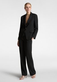 Abito nero su misura con giacca aderente caratterizzata da un singolo bottone dorato, pantaloni a gamba dritta e sandali con tacco alto e cinturini.