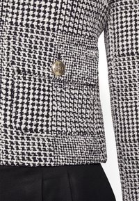 Vävd jacka med ett svart och vitt hundtandsmönster, med en guldmetallknapp och textil med struktur. Svart tyg synligt nedanför.