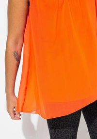 Top senza maniche arancione brillante realizzato in tessuto leggero, caratterizzato da una vestibilità ampia e sottili pieghe, abbinato a leggings neri a fantasia.