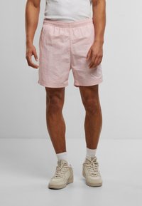 Lyse rosa shorts med elastisk liv, med teksturert stoff og et hvitt grafisk detalj på venstre ben. Brukt med kremfargede joggesko.