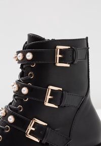 Bottine noire en cuir, avec quatre boucles dorées et des accents en perles. Surface texturée, bout arrondi et design à lacets.