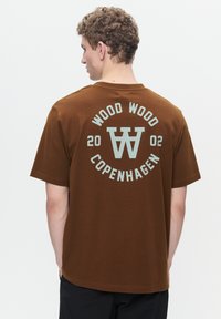 Brown cotton t-shirt with a crew neck. Back design features "WOOD WOOD COPENHAGEN" and a circular logo in light blue. Short sleeves, relaxed fit.