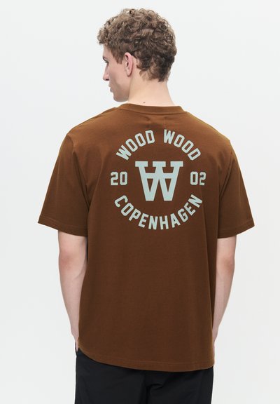 T-shirt di cotone marrone con collo rotondo. Il design sul retro presenta "WOOD WOOD COPENHAGEN" e un logo circolare in azzurro chiaro. Maniche corte, vestibilità comoda.