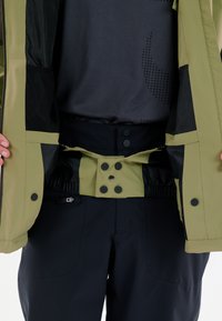 Oliven grøn jakke med sorte detaljer, som har lynlås og trykknaplukning, elastisk talje og indvendige mesh-lommer. Mørke bukser nedenfor.