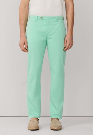 Hombre vistiendo pantalones chinos ajustados color verde menta con zapatos beige sin cordones, de pie contra un fondo liso de color gris claro.