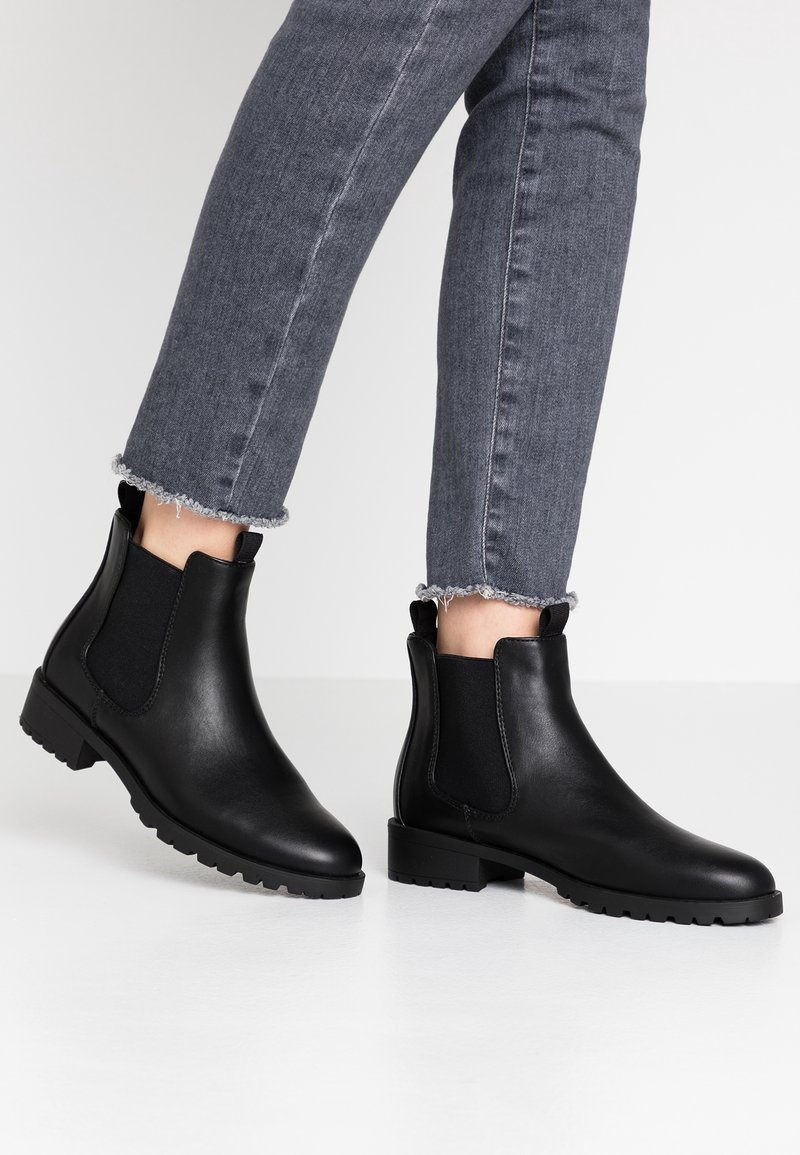 Bottines en cuir noir avec panneaux latéraux élastiques portées avec un jean en denim gris raccourci sur un sol blanc.