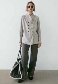 Maßgeschneidertes, zweireihiges gestreiftes Hemd in Hellgrau und Weiß, kombiniert mit weit geschnittenen, dunkelgrauen Hosen. Hält eine große, handschmeichlerische Handtasche.