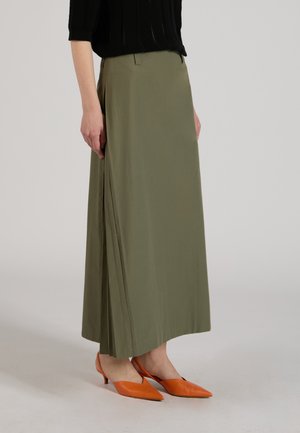 Osoba nosząca oliwkową plisowaną długą spódnicę w zestawieniu z pomarańczowymi czółenkami typu slingback z ostrym noskiem, stojąca na neutralnym tle.