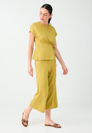 Woman in olive-green short-sleeve top with waist tie and matching wide-leg cropped pants, wearing brown flat sandals, standing on white floor.