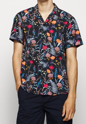 Mann trägt schwarzes kurzärmeliges Hemd mit buntem Blumenmuster und dunkle Hose, Hände in den Taschen.