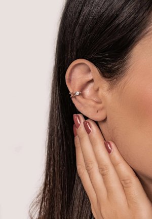 Pendiente de aro plateado con una sola piedra preciosa clara, usado en la parte superior de la oreja. Superficie de metal suave con un acabado pulido. Pelo corto visible.