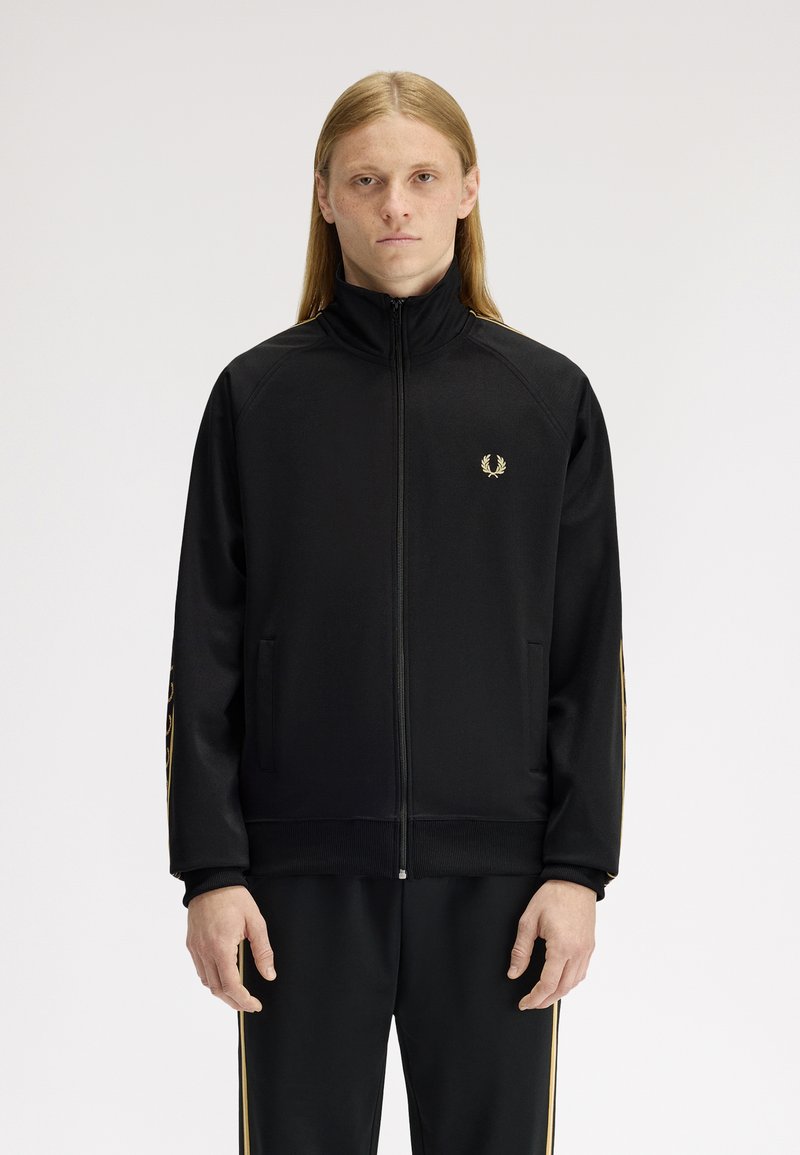 Giovane con capelli lunghi in piedi di fronte, indossa una giacca nera con zip e pantaloni coordinati con strisce dorate.