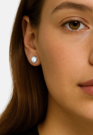Orecchino a bottone circolare in argento con una pietra centrale trasparente, finitura lucida, indossato nell'orecchio di una persona con capelli lunghi.