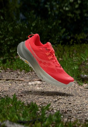 Chaussure de course rouge vif avec une semelle blanche épaisse flottant au-dessus d'un sentier de gravier entouré d'herbe verte et de feuillage.