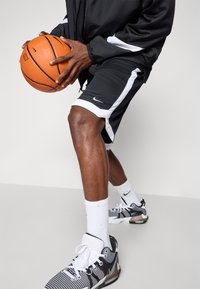 Pantalones cortos deportivos negros con franjas blancas, una chaqueta negra, calcetines blancos y zapatillas negras/blancas con un patrón, sosteniendo un balón de baloncesto naranja.