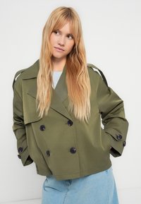 Oliivinvihreä, lyhyt trenssitakki, jossa kaksirivinen napitus, mustat napit ja olkasaappaat. Päällä vaaleansinisen topin päällä.