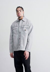 Light gray denim jacket with a collar, two chest pockets, and button closure. Worn over black pants against a white background.