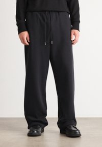 Schwarze Sweatpants aus weichem Stoff, mit einem elastischen Bund mit Kordelzug und einer lockeren Passform. Kombiniert mit schwarzen Schuhen.