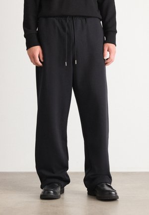 Zwarte sweatpants gemaakt van zacht materiaal, met een elastische tailleband en trekkoord, in een relaxte pasvorm. Gecombineerd met zwarte schoenen.