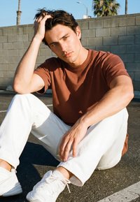 Młody mężczyzna o ciemnych włosach, ubrany w brązową koszulę i białe spodnie, siedzi na asfalcie obok ceglanej ściany i palm, opierając rękę na głowie.