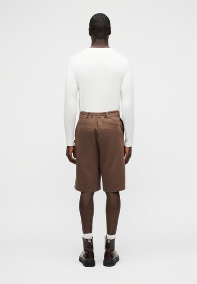 Άτομο που στέκεται με την πλάτη στραμμένη φορώντας λευκό μακρυμάνικο μπλουζάκι, καφέ σορτς μέχρι το γόνατο, λευκές κάλτσες και σκούρα καφέ μπότες σε απλό φόντο.