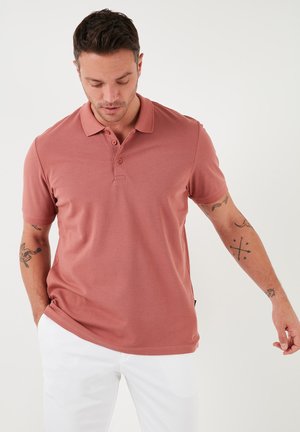 Polo de manga corta en una mezcla de algodón en rosa empolvado, con cuello y tapeta de tres botones. Cuenta con una abertura lateral para mayor comodidad.