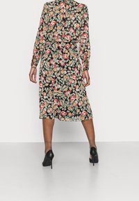 Robe fleurie noire à manches longues, avec un motif coloré de fleurs, une taille cintrée et un ourlet arrivant au genou. Associée à des talons noirs.