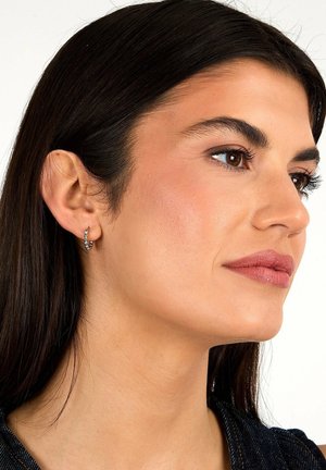 Woman with straight dark hair wears small silver hoop earrings, looking slightly to the right against a plain white background.