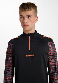 Svart sporttopp med långa ärmar. Har orange dragkedjor och logotyp. Ärmarna har ett mönstrat textur i nyanser av röd och svart.