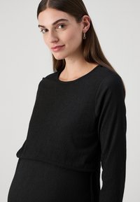 Crna majica dugih rukava s teksturiranom tkaninom, okruglim izrezom i slojevitim dizajnom donjeg dijela. Mekan materijal, prilagođen oblik i minimalni detalji.