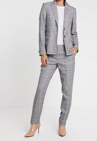 Kobieta ubrana w szary garnitur w kratę z dopasowaną marynarką i pasującymi spodniami, białą bluzkę oraz nude lakierowane botki na wysokim obcasie.