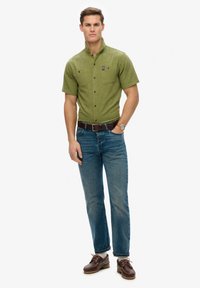 Camisa de manga corta, color verde oliva, con botones y dos bolsillos en el pecho, combinada con jeans azules y zapatos marrones, complementada con un cinturón marrón.