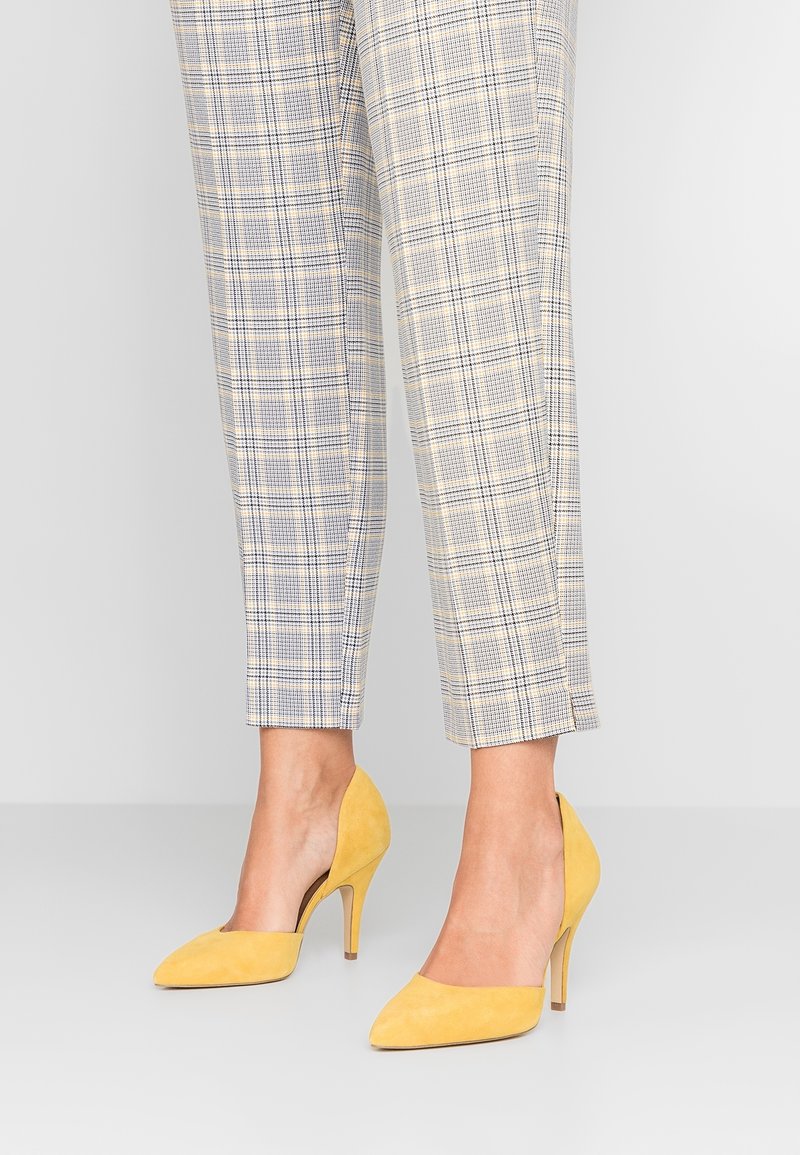 Yellow suede high-heeled shoes with a pointed toe and cut-out sides, worn with light plaid tailored pants featuring blue and beige patterns.