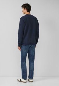 Granatowa teksturowana bluza z prążkowanymi mankietami, zestawiona z niebieskimi jeansami o prostym kroju oraz białymi trampkami z czarnymi akcentami. Widok od tyłu.