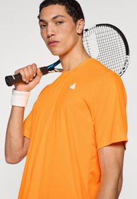 Φωτεινό πορτοκαλί αθλητικό μπλουζάκι με κοντά μανίκια, κατασκευασμένο από ελαφρύ ύφασμα. Μαύρη ρακέτα τένις κρατημένη σε ένα χέρι, με περιβραχιόνιο στον καρπό.