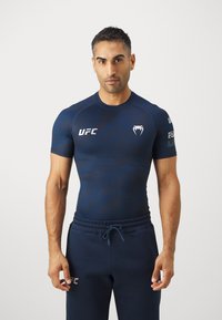 Jūrinis sportinis marškinėliai su trumpomis rankovėmis, puikuojasi UFC logotipu ir tekstūruotu dizainu. Suderinti su jūrinėmis sportinėmis kelnėmis, kurios demonstruoja laisvą stilių.