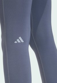 Grijze sportlegging met een gladde textuur, voorzien van contrasterende lichtgrijze stiksels en een wit Adidas-logo op de linker dij.