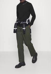 Man som går i en svart Tommy Hilfiger långärmad tröja, olivgröna byxor, svarta stövlar, med en svartvit rutig tröja knuten runt midjan.