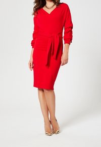 Rotes, figurbetontes Kleid mit Wickeldesign, V-Ausschnitt, langen Ärmeln und gebundenem Taillendetail. Getragen mit beigen High Heels.