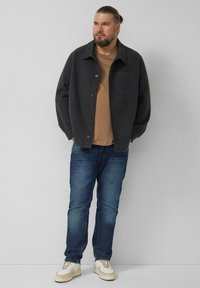 Ciemnoszara marynarka z mieszanki wełny z kołnierzem, przednimi guzikami i pojedynczą kieszenią na piersi, noszona na brązowej koszuli oraz niebieskich dżinsach, do tego kremowe trampki.