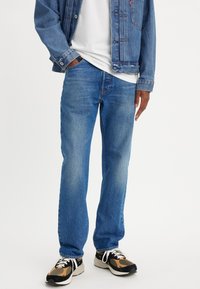 Jean en denim bleu avec une coupe droite, délavage moyen et légère usure, associés à un t-shirt blanc et des baskets à motif.