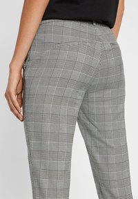 Kvinna i grå rutiga byxor med en sidoficka och en svart topp, hand avslappnad vid sidan.