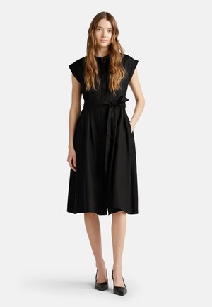 Schwarzes Leinenkleid mit kurzen Ärmeln, Knopfverschluss vorne, Gürtelschnürung in der Taille und knielanger Rock. Kombiniert mit spitzen schwarzen High Heels.