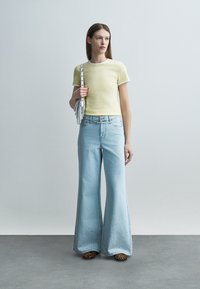 Kobieta w żółtej bluzce z krótkim rękawem i szerokich jasnoniebieskich dżinsach z ozdobnymi wykończeniami przy nogawkach, trzymająca białą torebkę, stojąca na szarej podłodze.