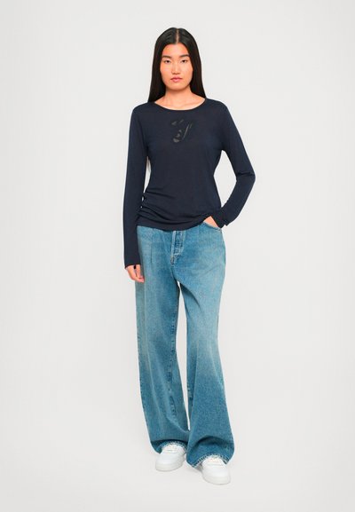 Mlada žena s dugom tamnom kosom nosi tamnoplavu majicu dugih rukava, široke svijetloplave traperice i bijele tenisice, stoji pred jednostavnom pozadinom.