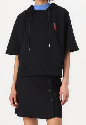 Person trägt einen schwarzen Hoodie mit kurzen Ärmeln und einem roten Chili-Patch sowie einen schwarzen Rock mit drei vorderen Knöpfen über einem blauen Rollkragenpullover.